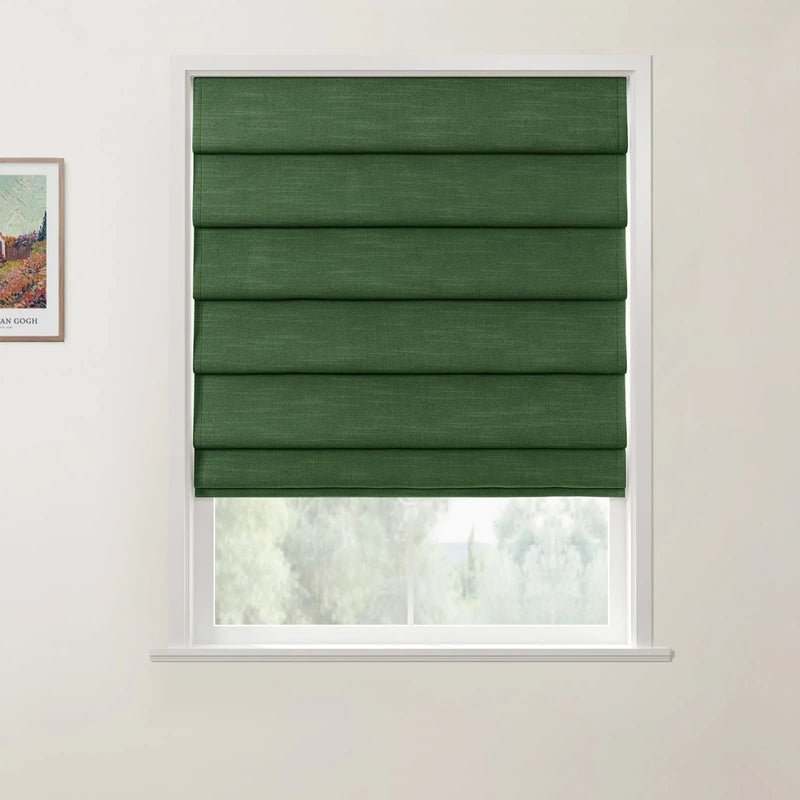 A photograph showcasing a window with a dark green textured fabric Roman blind partially raised above a view of blurred green trees, next to a framed print of a colorful Van Gogh landscape painting on a white wall