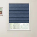 A dark blue fabric Roman blind with neatly folded horizontal pleats covering a white framed window, with a partial Van Gogh print visible on the wall and blurred outdoor greenery