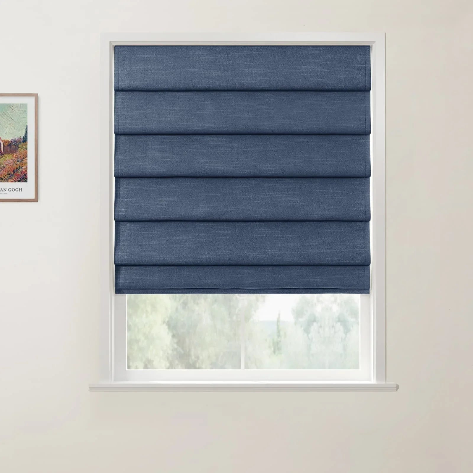 A dark blue fabric Roman blind with neatly folded horizontal pleats covering a white framed window, with a partial Van Gogh print visible on the wall and blurred outdoor greenery