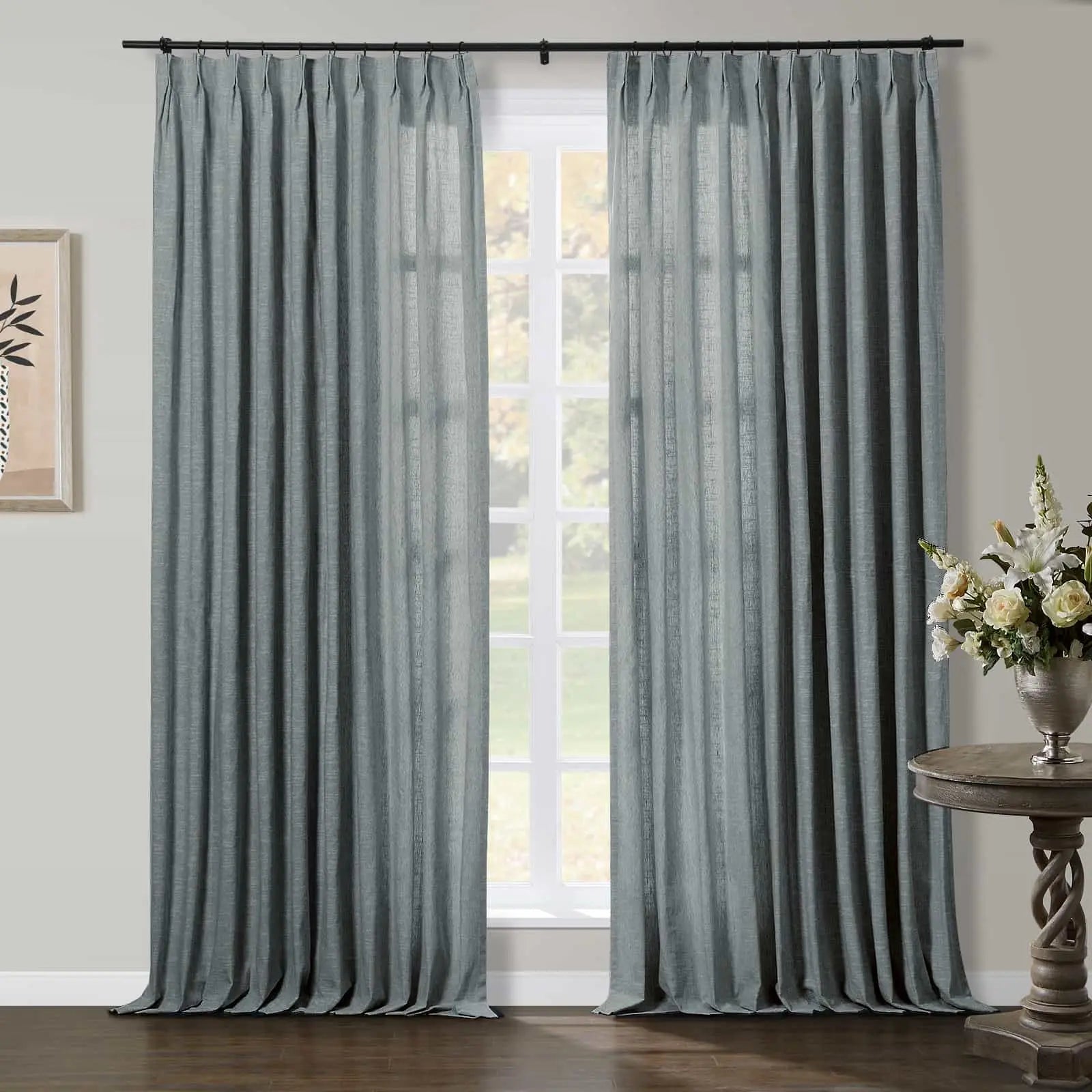 Photograph of a room interior featuring elegant floor-length blue-grey linen pinch pleat curtains flanking a window with natural light and blurred green trees, accented by a white floral arrangement on a carved wooden side table