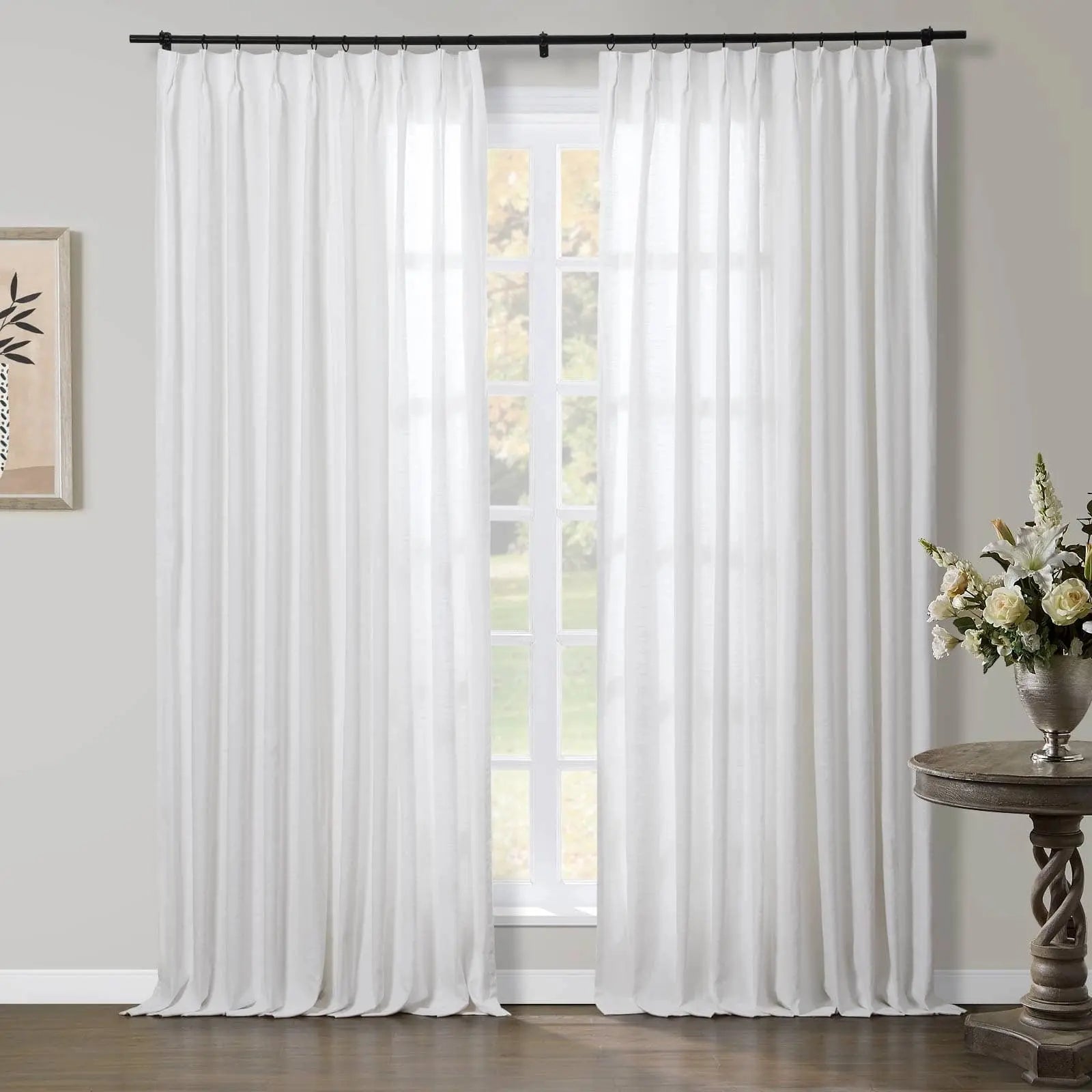 Photograph of sheer white pinch-pleat curtains on a black rod in a bright room with a multi-pane window overlooking a lush green outdoor view, featuring a framed botanical print and an ornate wooden side table holding a silver vase of white and cream flowers