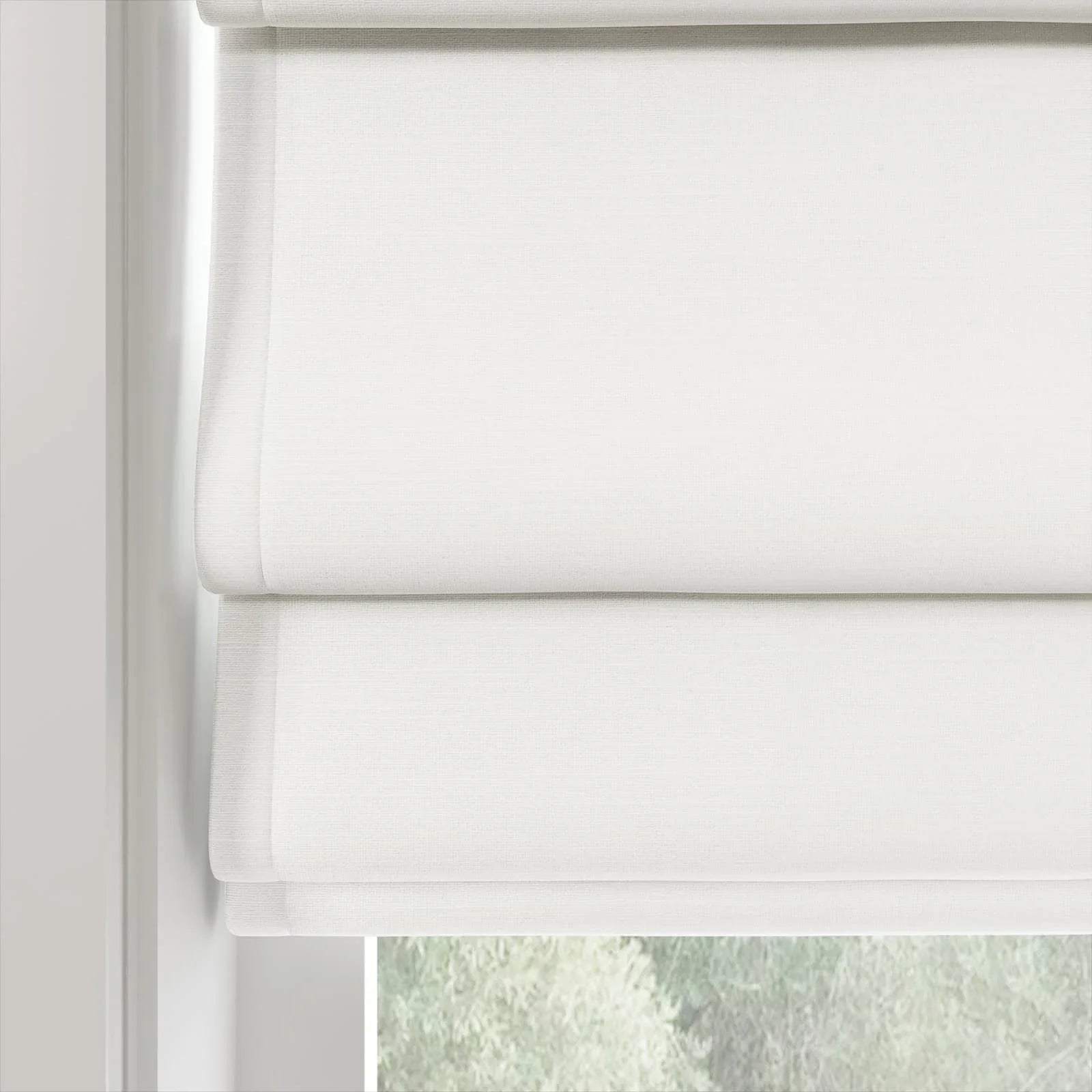 White textured fabric Roman blinds partially open at a window, revealing blurred green foliage outside
