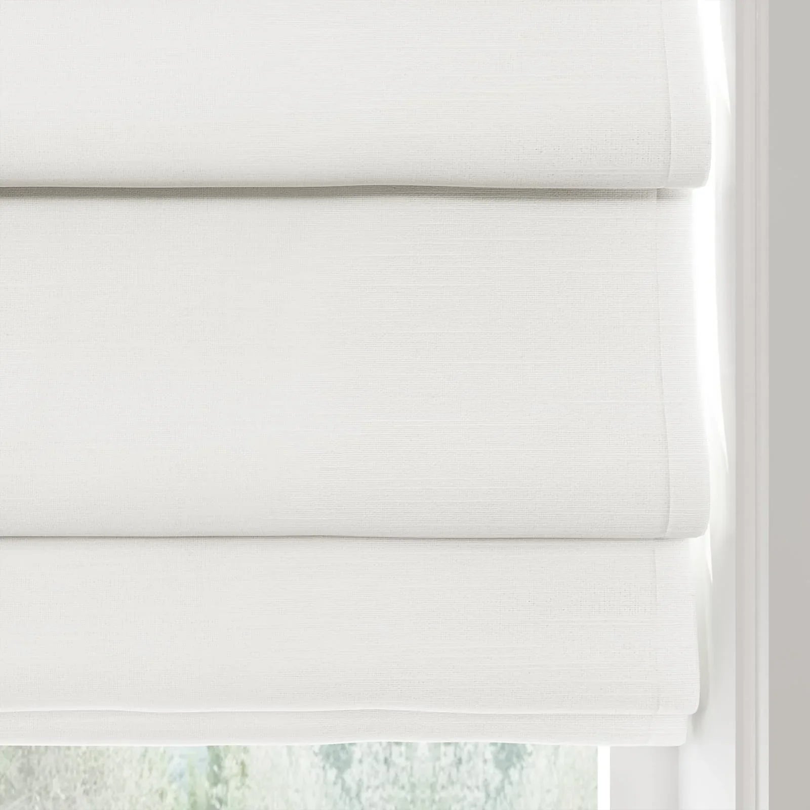 White textured Roman blinds featuring three clean horizontal folds partially covering a window, softly lit by natural light with a hint of blurred green trees beyond