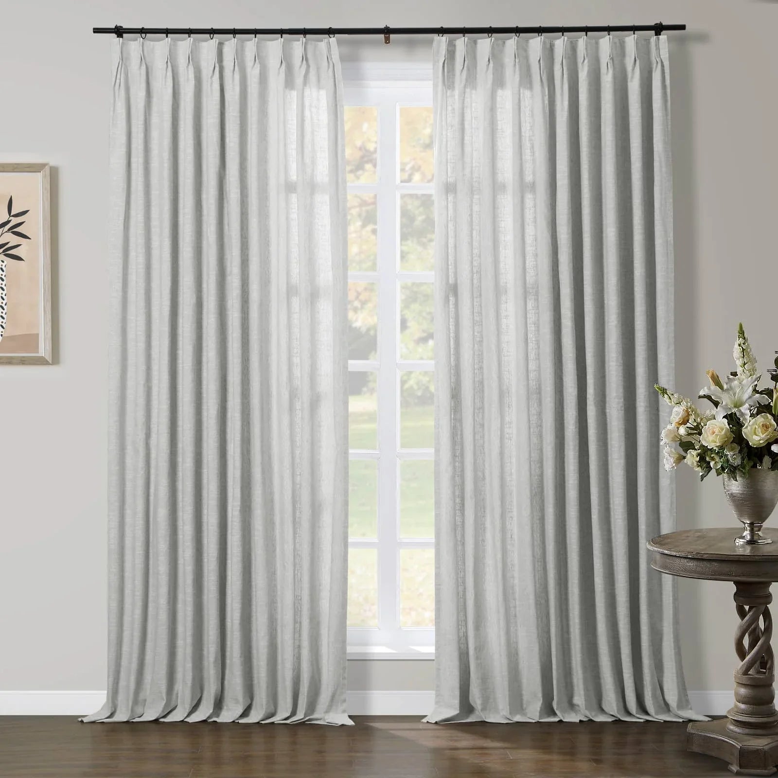 Light grey linen-look pinch pleat curtains on a black rod framing a large window, complemented by a decorative wooden side table with a silver vase of white and yellow flowers and abstract botanical framed art on a muted grey wall