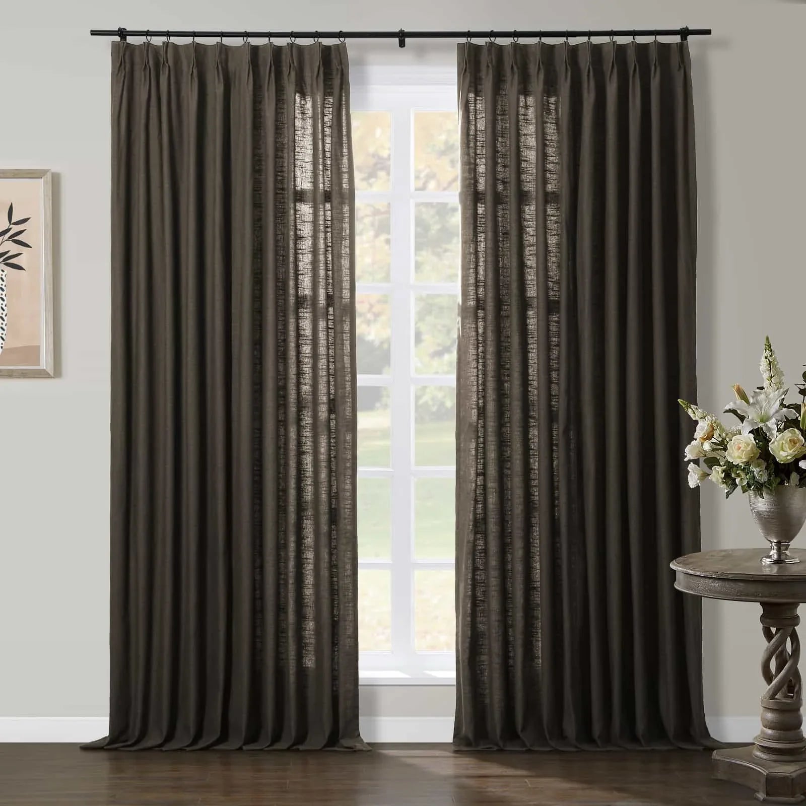 Dark brown textured linen curtains hang on a black rod framing a white-paned window with an outdoor greenery view, complemented by a minimalist framed plant artwork and a silver vase of white roses and lilies on a carved wooden side table