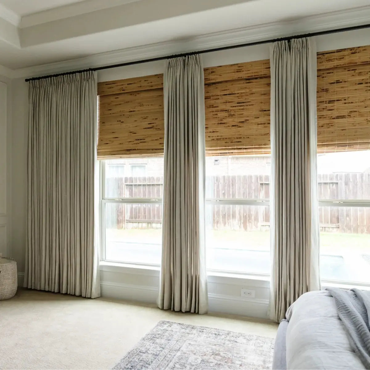 Modern bedroom interior featuring four large windows with natural woven wood shades and elegant beige pinch-pleat drapes on black rods