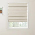 Photograph of a window featuring a cream textured fabric Roman blind with neat horizontal folds, partially revealing blurred green trees outside, with a framed Van Gogh art print on the adjacent white wall