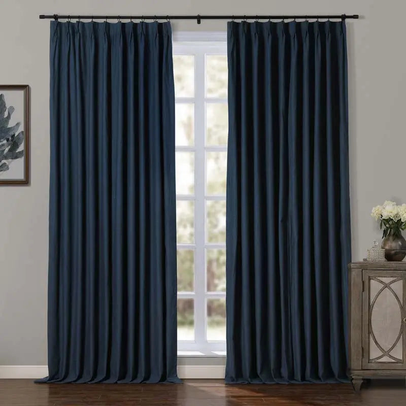 A pair of navy blue pinch pleat curtains hang floor-length from a black rod in a bright room, alongside a distressed wooden cabinet with white flowers and a framed abstract print