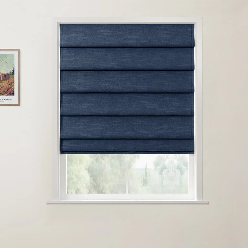 Photograph showing a dark blue textured Roman blind over a white-framed window, with blurred green foliage visible below and a partial framed Van Gogh landscape print on the adjacent light wall