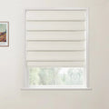 Photograph of a white-framed window featuring neatly stacked cream Roman blinds in an interior room, with a partially visible Van Gogh print on the left wall and blurred green foliage outside