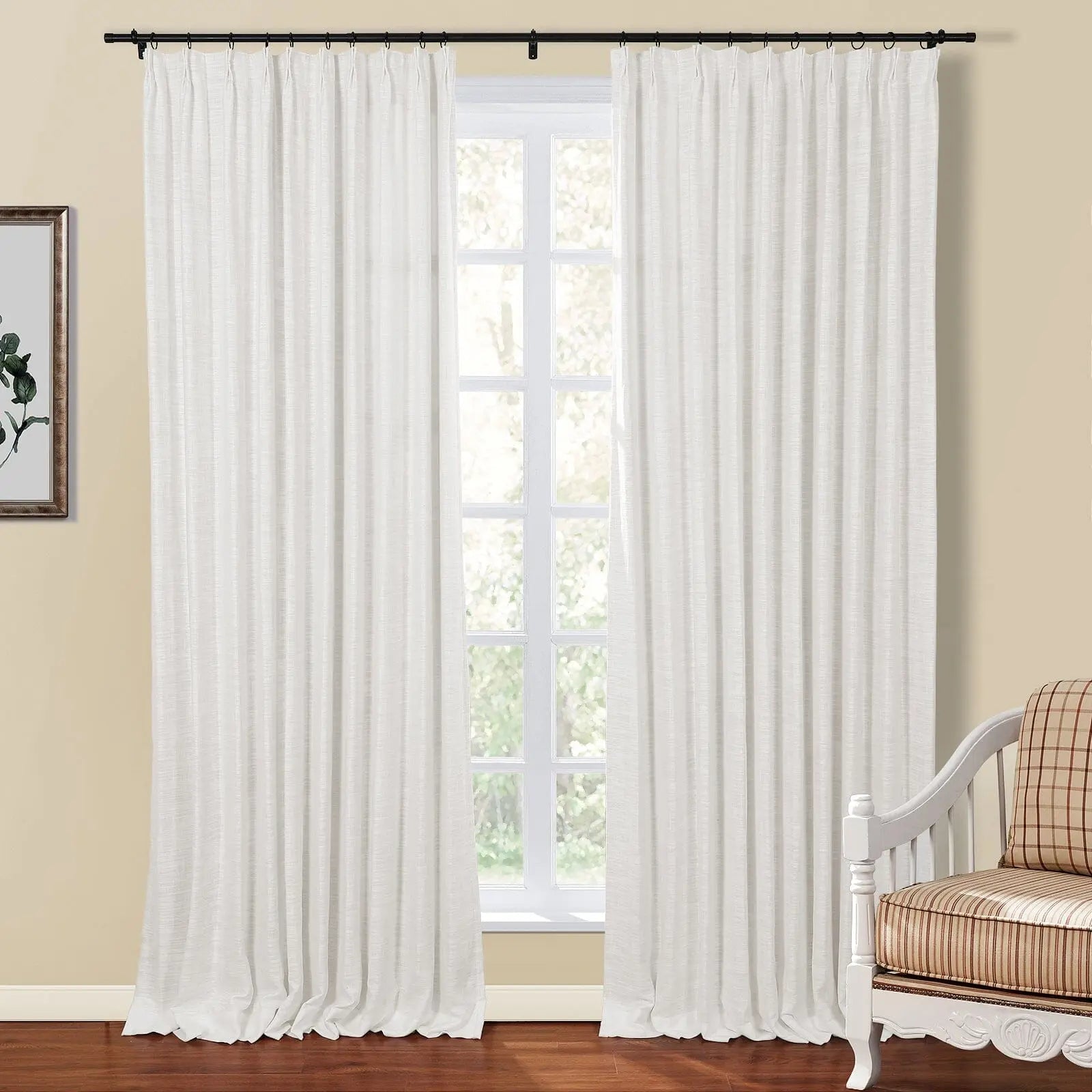 A bright room with a large window featuring white textured linen-look curtains on a black rod, revealing vibrant green foliage outside, complemented by a framed botanical print on the left wall and a plaid and striped cushioned armchair on the right