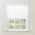 A white Roman shade covers the top half of a multi-pane window looking out onto a blurry green yard, with a white wall-mounted remote control on the adjacent beige wall