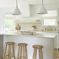 A modern minimalist kitchen with light beige marble countertops and backsplash, three natural wood bar stools, white cone pendant lights, and a prominent brass faucet