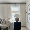 Interior photograph of a cozy living room featuring a cream sofa with striped throw pillows, layered white window treatments overlooking a snowy landscape, a rustic terracotta lamp on a black end table, and a slender brass wall sconce