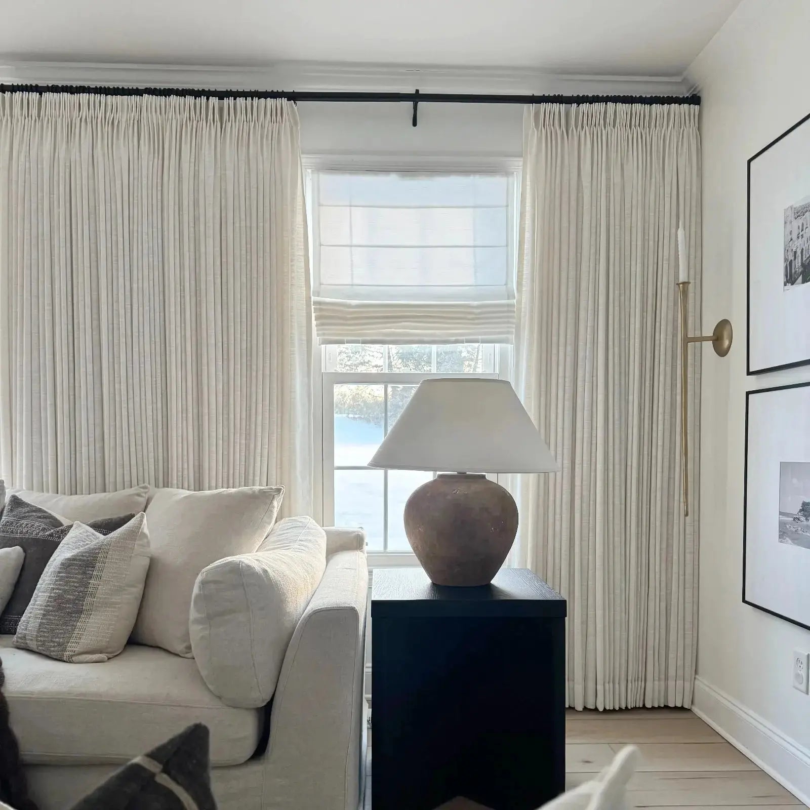 Interior photograph of a cozy living room featuring a cream sofa with striped throw pillows, layered white window treatments overlooking a snowy landscape, a rustic terracotta lamp on a black end table, and a slender brass wall sconce
