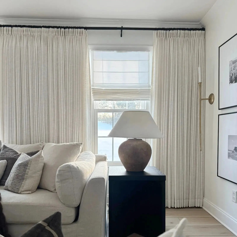 Interior photograph of a cozy living room featuring a cream sofa with striped throw pillows, layered white window treatments overlooking a snowy landscape, a rustic terracotta lamp on a black end table, and a slender brass wall sconce