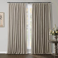 Floor-length natural linen pleated drapery panels on a black rod frame a white gridded window in a modern room with a dark wood floor and a decorative side table holding a white floral arrangement