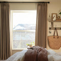 A cozy bedroom interior with beige drapes framing a window overlooking a snowy roof, featuring a woven straw tote bag hanging on the wall and a knitted rust-colored throw blanket on the bed