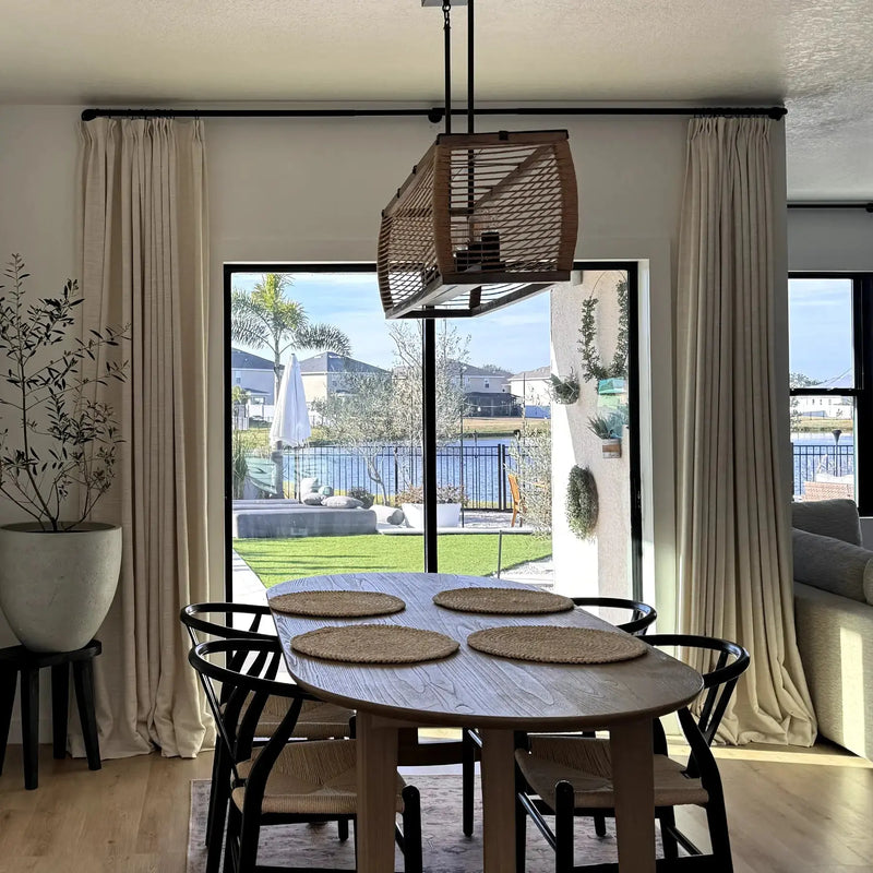 Photograph of a modern dining room featuring an oval wood table with woven placemats and black Wishbone chairs, illuminated by a distinctive rectangular woven chandelier, offering a sunlit view of a lush green backyard, concrete patio seating, a white outdoor umbrella, and a calm lake with distant houses and palm trees
