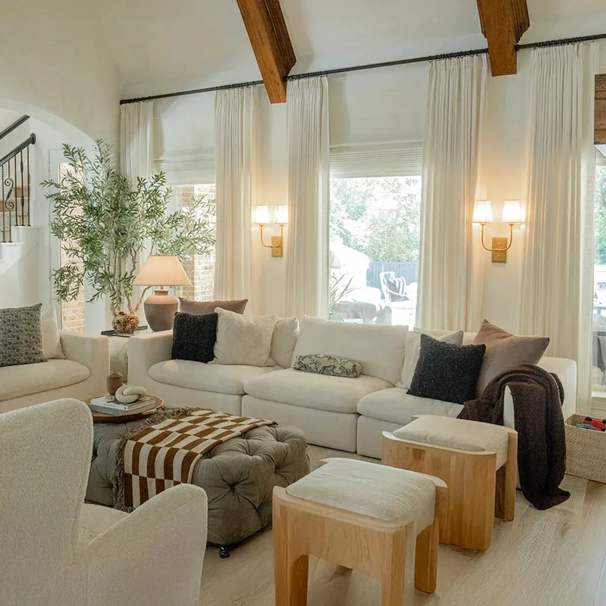 A modern living room interior featuring a white modular sofa exposed dark wood ceiling beams sheer white curtains with brass wall sconces a tufted ottoman adorned with a brown and white checkered throw and a large potted olive tree