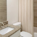Photograph of a modern bathroom interior featuring a beige granite countertop with an undermount sink and brushed nickel faucet, a white toilet, and a pleated cream shower curtain beside a light brown tiled shower wall