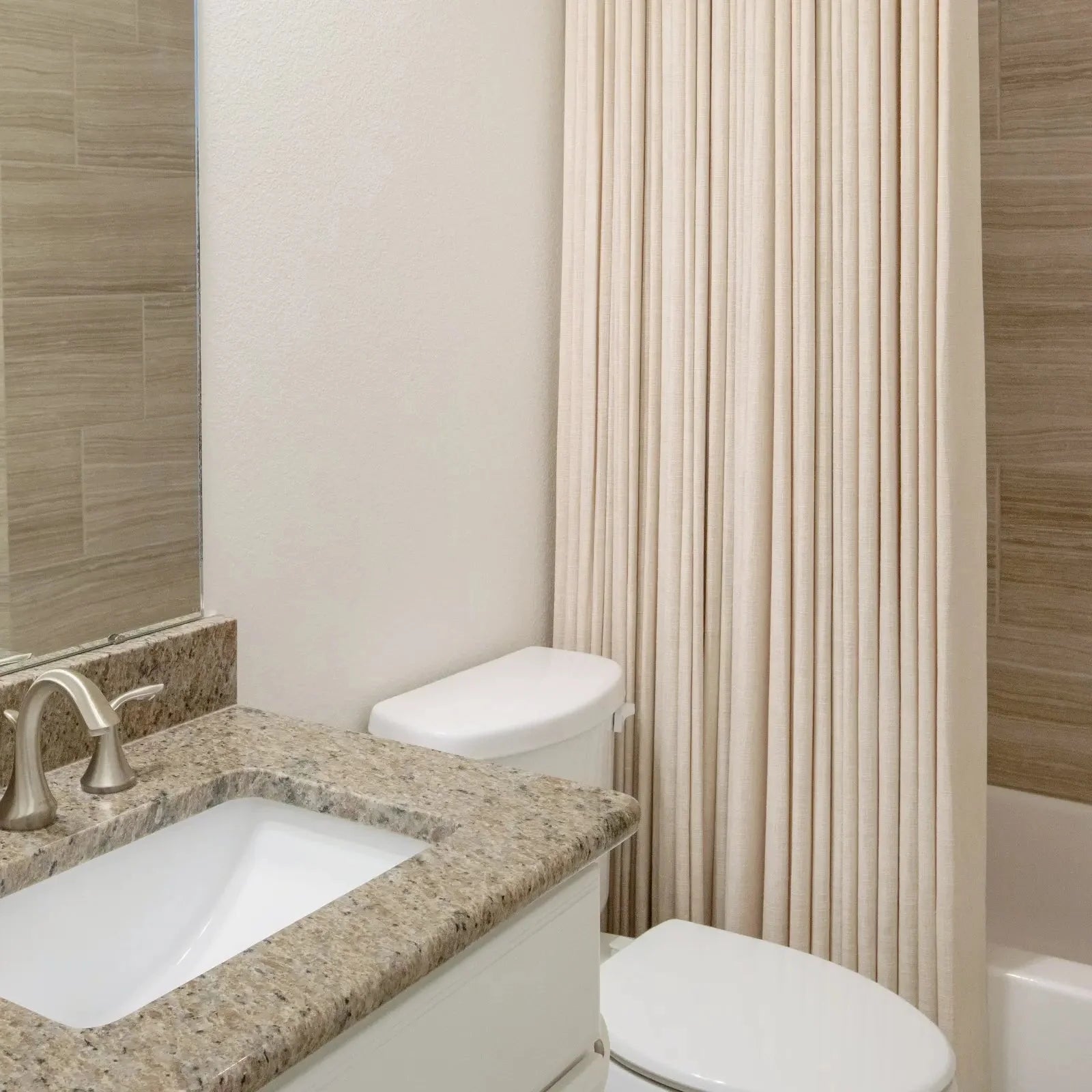 Photograph of a modern bathroom interior featuring a beige granite countertop with an undermount sink and brushed nickel faucet, a white toilet, and a pleated cream shower curtain beside a light brown tiled shower wall