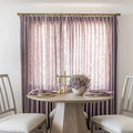 A stylish dining area in an interior room featuring pale lavender textured drapes a round light wood table with a geometric base gold candlesticks and purple foliage in a white vase