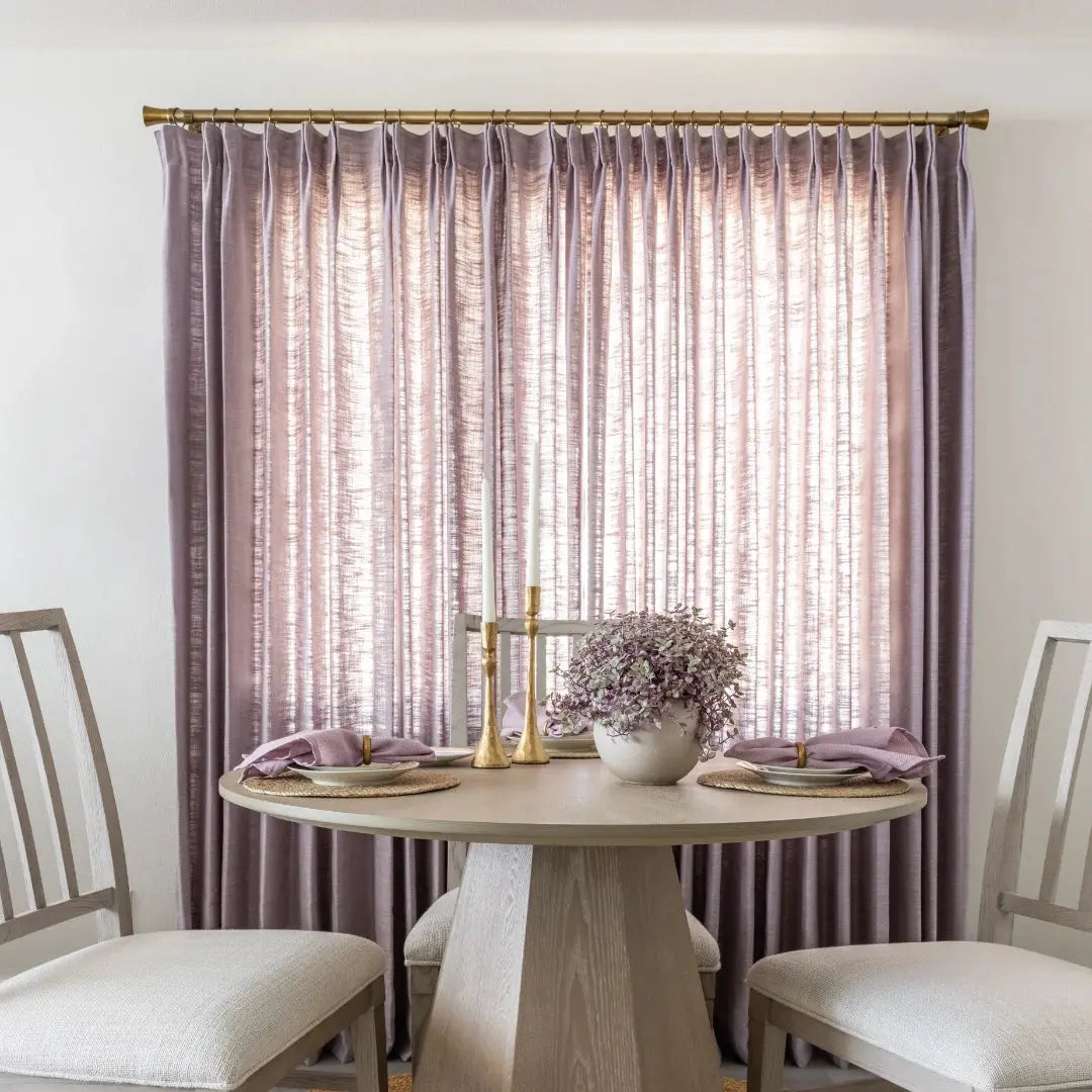 A stylish dining area in an interior room featuring pale lavender textured drapes a round light wood table with a geometric base gold candlesticks and purple foliage in a white vase