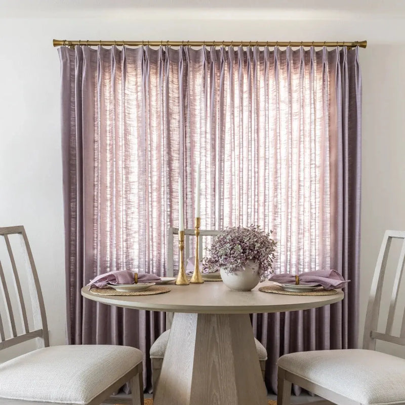 A stylish dining area in an interior room featuring pale lavender textured drapes a round light wood table with a geometric base gold candlesticks and purple foliage in a white vase