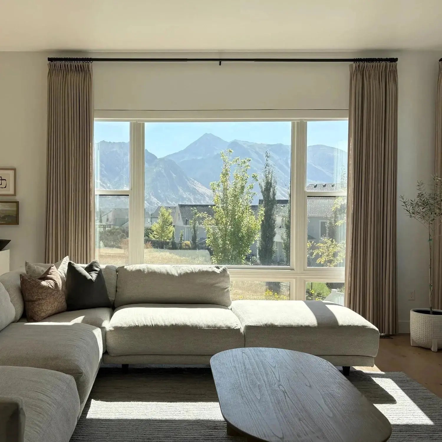 Photograph, unknown artist, a modern living room with a light grey sectional sofa and oval wooden coffee table, brightly lit by sunlight streaming through a large window overlooking a distant mountain range and green trees