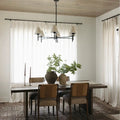A modern rustic dining room interior featuring a dark wood dining table with woven-back chairs, a centerpiece of two rustic vases with vibrant green foliage, and a sleek black chandelier with cream shades under a light wood planked ceiling