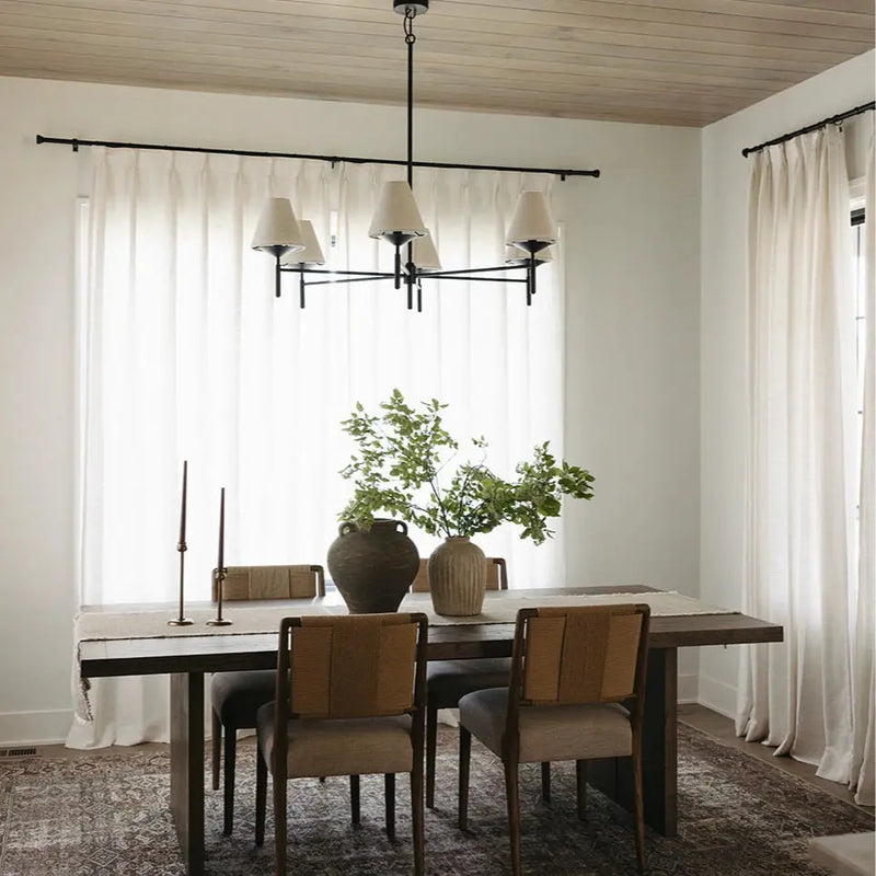 A modern rustic dining room interior featuring a dark wood dining table with woven-back chairs, a centerpiece of two rustic vases with vibrant green foliage, and a sleek black chandelier with cream shades under a light wood planked ceiling