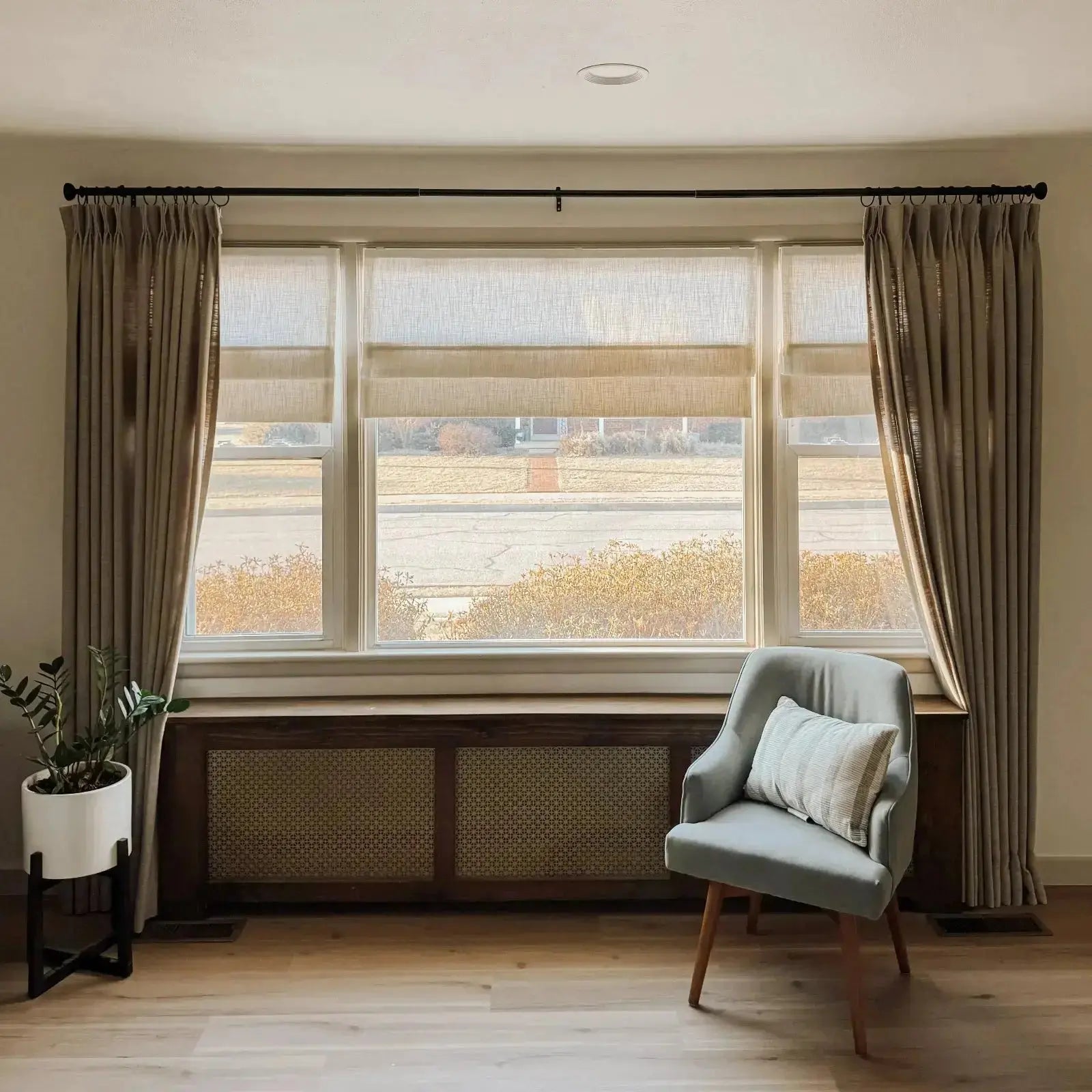 Interior view of a living room featuring a large triple window with light beige Roman shades and drapes, a dark wood radiator cover with rattan cane panels, a light gray accent chair with a striped pillow, and a potted plant on light wood flooring