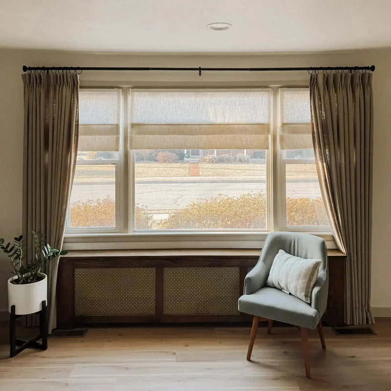 Interior view of a living room featuring a large triple window with light beige Roman shades and drapes, a dark wood radiator cover with rattan cane panels, a light gray accent chair with a striped pillow, and a potted plant on light wood flooring