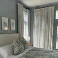 Photograph of a serene bedroom interior featuring gray-blue walls a cream upholstered headboard gray-green toile floral bedding two framed botanical prints and floor-length cream curtains