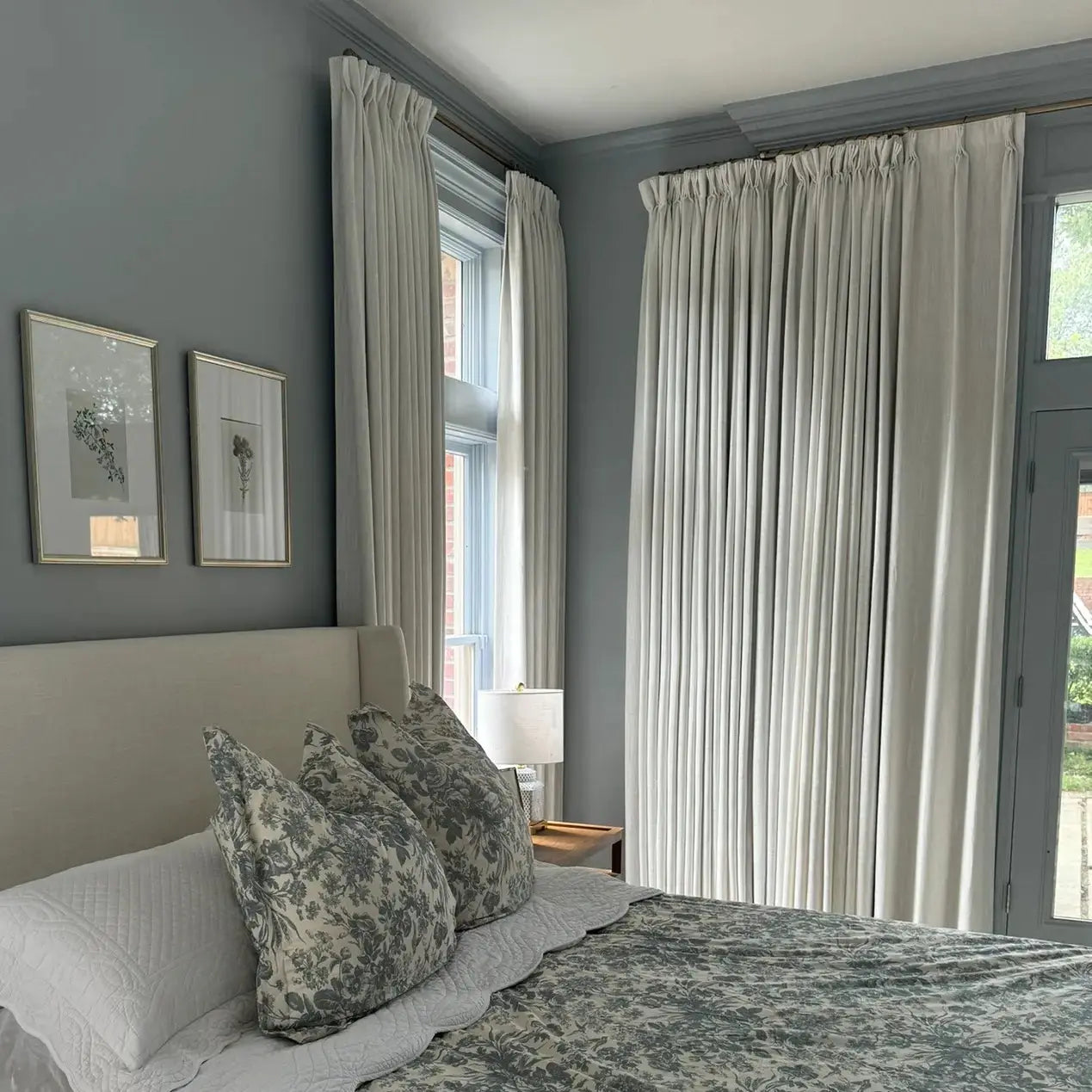 Photograph of a serene bedroom interior featuring gray-blue walls a cream upholstered headboard gray-green toile floral bedding two framed botanical prints and floor-length cream curtains