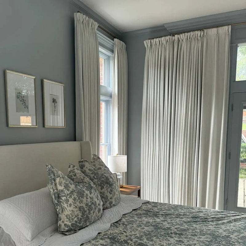 Photograph of a serene bedroom interior featuring gray-blue walls a cream upholstered headboard gray-green toile floral bedding two framed botanical prints and floor-length cream curtains