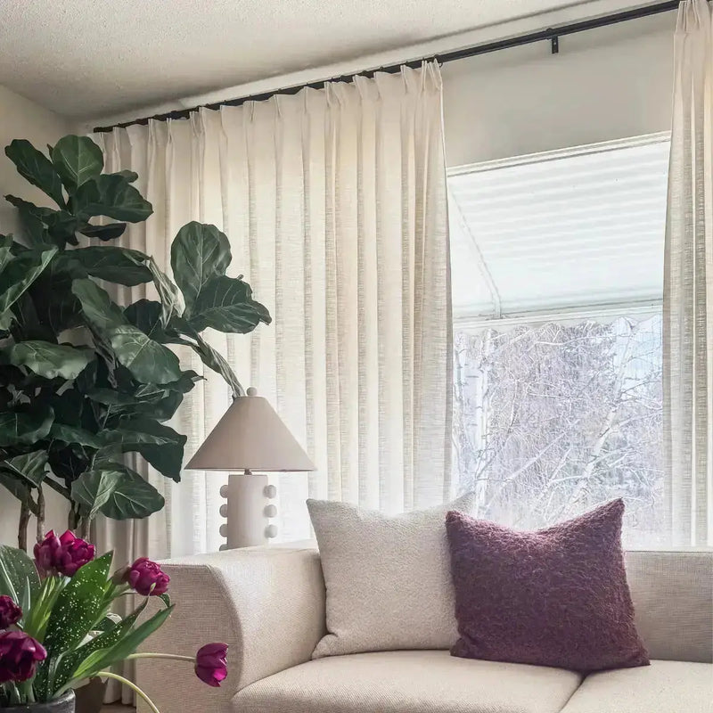 A stylish living room with a large fiddle-leaf fig tree, a beige sofa with a dark purple fuzzy pillow, and vibrant purple tulips on a side table overlooking a snowy winter scene