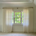 Bright interior room with off-white pleated linen-like curtains on a brass rod framing a grid-paned wooden window revealing a vibrant green tree canopy outside and featuring white crown molding and light neutral carpet