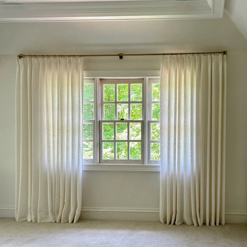 Bright interior room with off-white pleated linen-like curtains on a brass rod framing a grid-paned wooden window revealing a vibrant green tree canopy outside and featuring white crown molding and light neutral carpet