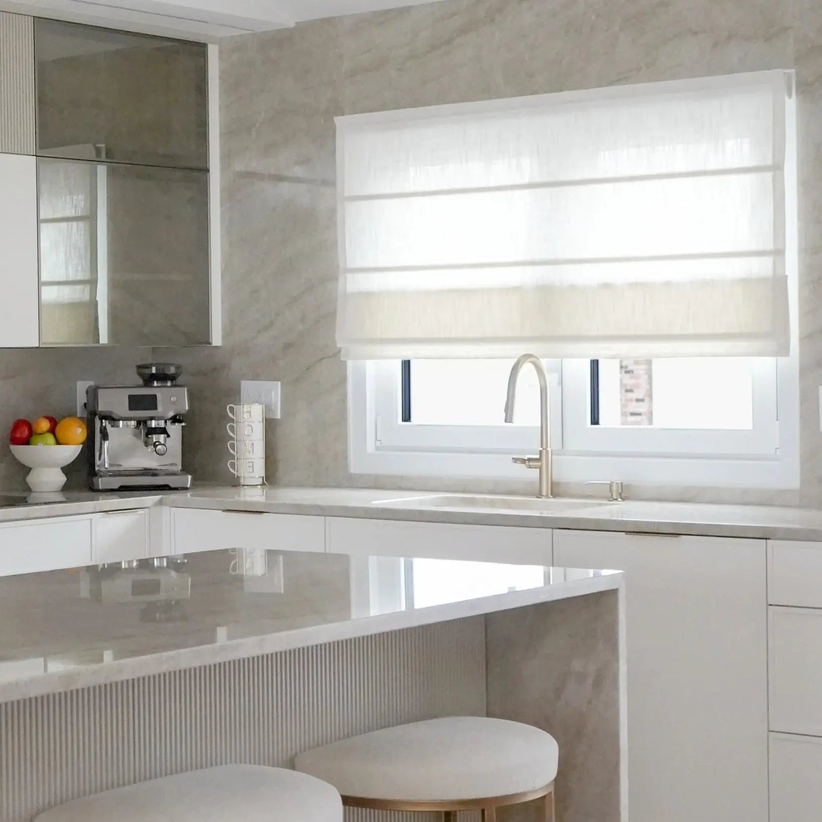 Photograph of a modern residential kitchen with light gray marble countertops, a sleek silver espresso machine, and a bowl of colorful fruit