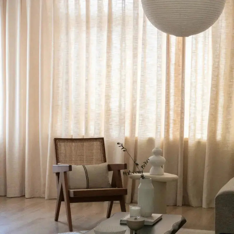 Modern minimalist living room interior with flowing beige linen curtains, a mid-century wooden armchair featuring a cane back and striped lumbar pillow, a white sculptural vase on a side table, and a large spherical paper lantern overhead