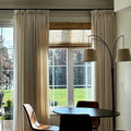 An interior dining nook with elegant beige floor-length curtains, a textured woven wood roman shade, a modern black two-armed floor lamp with beige shades, a dark oval dining table, and sleek wooden chairs overlooking a lush green backyard