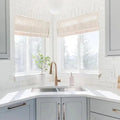Modern kitchen corner with a bright window over a double sink, featuring white painted brick walls, pale blue shaker cabinets with gold hardware, a brass gooseneck faucet, and a small green potted plant