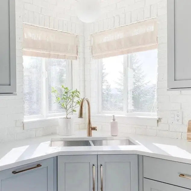 Modern kitchen corner with a bright window over a double sink, featuring white painted brick walls, pale blue shaker cabinets with gold hardware, a brass gooseneck faucet, and a small green potted plant