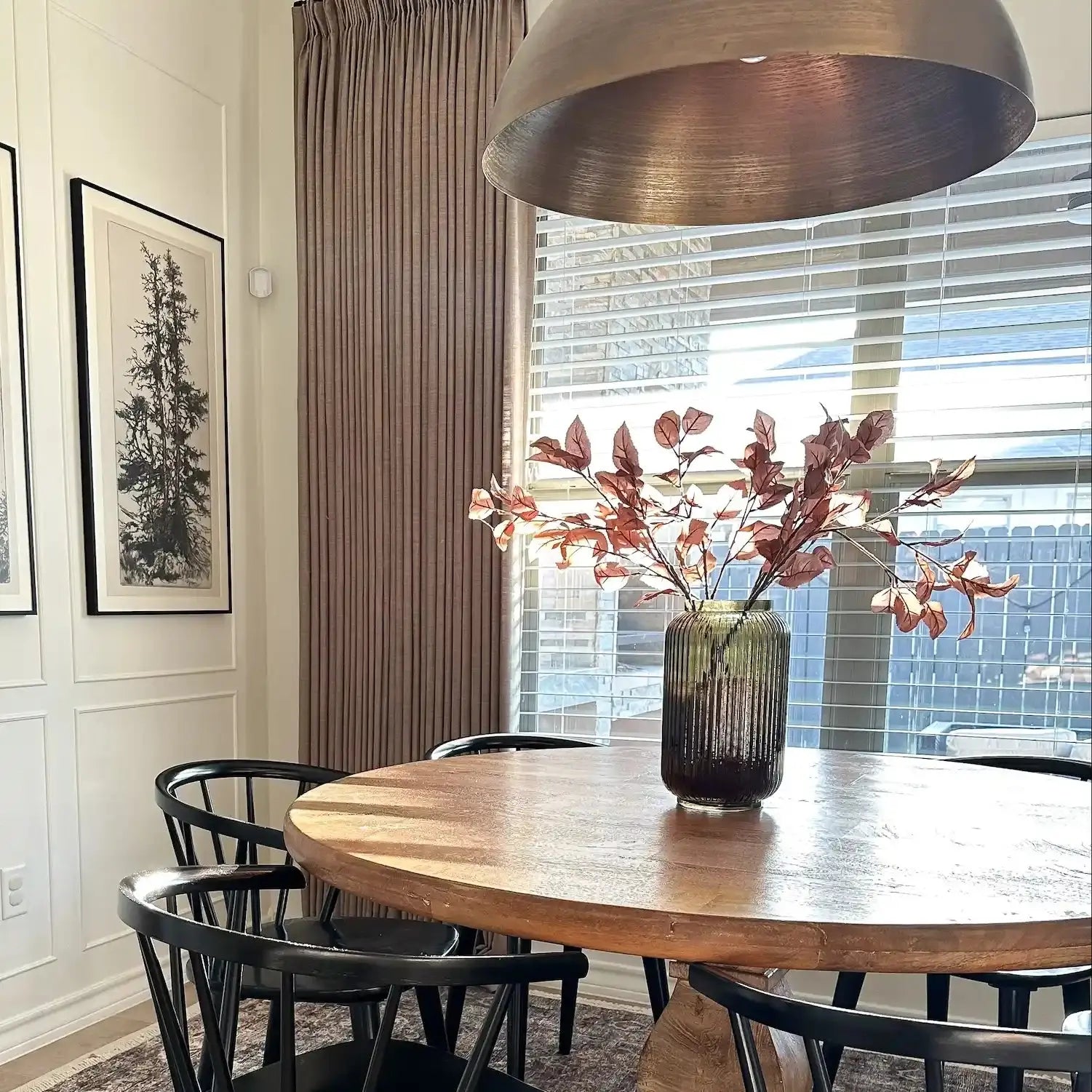 A modern dining room interior featuring a rustic round wood table, black Windsor chairs, a large bronze dome pendant light, and a fluted glass vase with vibrant rust-colored fall foliage