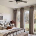 A cozy bedroom features a black metal bed frame with a terracotta velvet pumpkin, a nightstand holding dark red dried florals, and large windows with taupe drapes on a gold rod overlooking a green lawn and trees