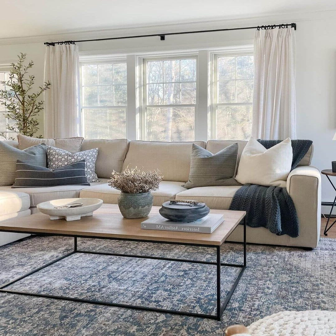 A bright modern living room showcases a neutral beige sectional sofa with various grey and white accent pillows, a stylish light wood and black metal coffee table with dried botanicals, and a detailed blue and white patterned area rug under large windows with sheer white curtains