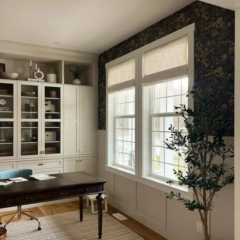 A modern home office interior featuring a dark floral wallpaper accent wall, white built-in cabinetry with brass hardware, a large dark wood desk with a light blue office chair, and a tall faux olive tree next to bright windows