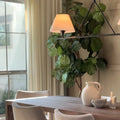 A bright photograph of a modern dining room featuring a prominent fiddle leaf fig tree, a linear chandelier with two illuminated lampshades, and a textured white ceramic pitcher with small bowls on a dark wooden table near a sunlit window